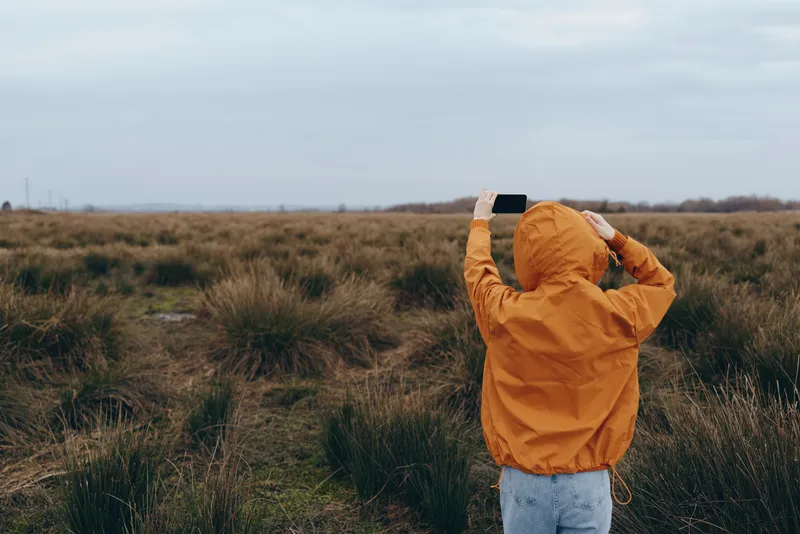 Woman in orange jacket walking through grassy field under cloudy sky, outdoor nature lifestyle in countryside, autumn breeze feeling, relaxed posture, woman lifestyle concept, outdoors