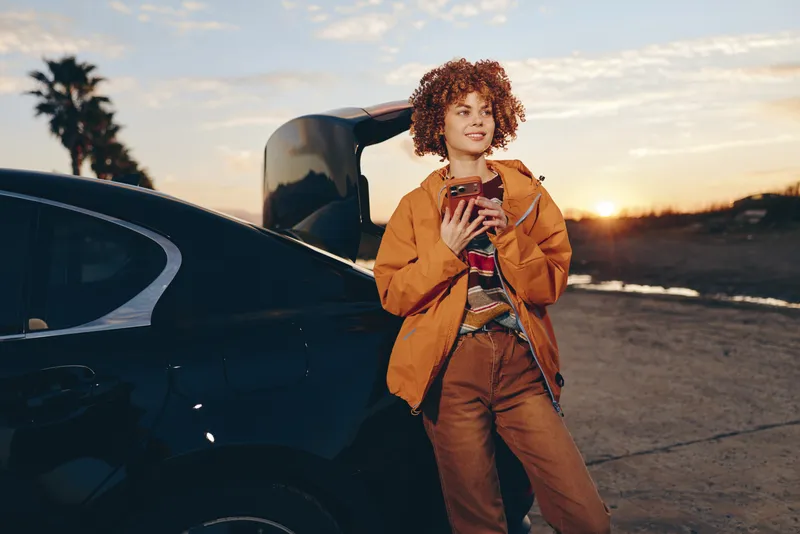 Woman lifestyle outdoors near car at sunset, curly hair, orange jacket, smiling and holding a coffee cup, relaxed stance by the roadside, warm golden light, scenic sky