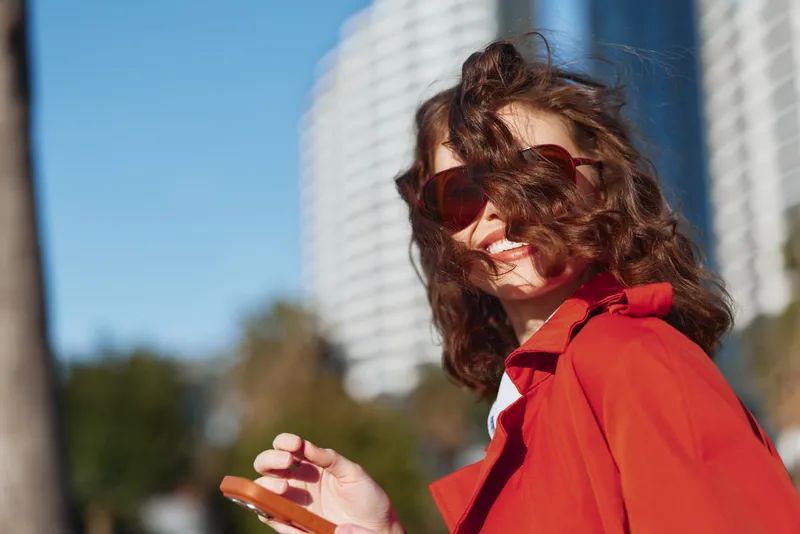 Young woman in a red jacket laughs outdoors with wind in her hair, sunglasses on, urban skyline behind, a carefree sunlit moment