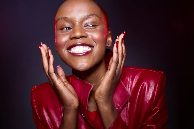 Smiling African woman with dark skin and shaved head wearing a red leather jacket poses happily against a dark background. Concept of beauty, confidence, fashion, and joy.