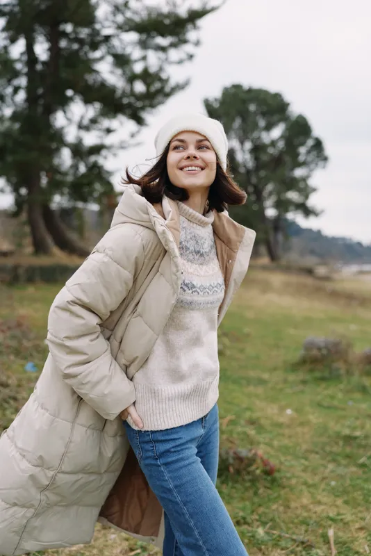 Woman outdoor winter coat smiling in a park, wearing a knit sweater and beanie, enjoying a breezy day with cozy warmth and natural light.
