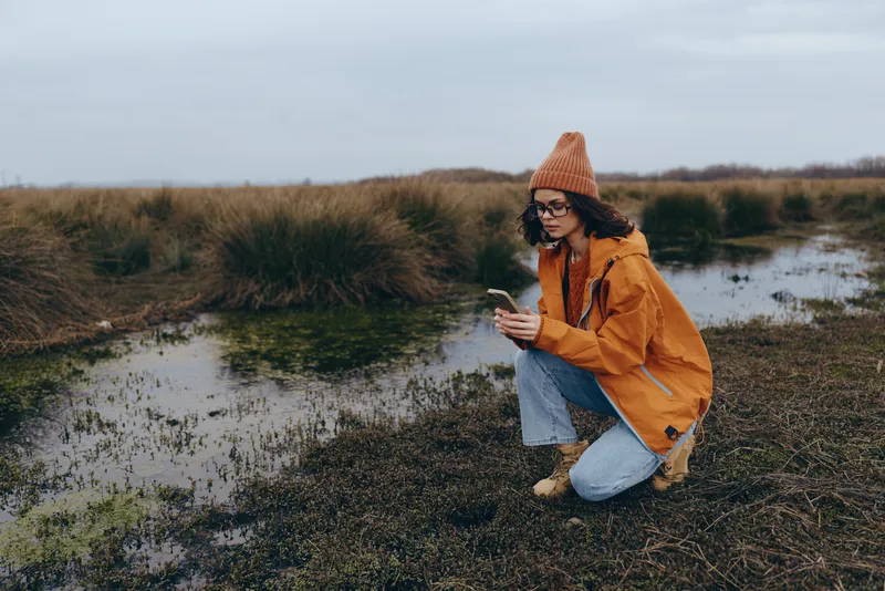 Woman lifestyle kneeling by riverbank, outdoor nature scene, wearing knit hat and orange jacket, holding phone with both hands, calm look toward water, cloudy sky and grass wetlands behind