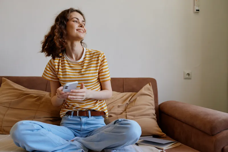 A relaxed woman sits on a couch at home, smiling while holding a game controller. Bright, casual setting shows daily leisure and comfortable living, with soft textiles and warm light.