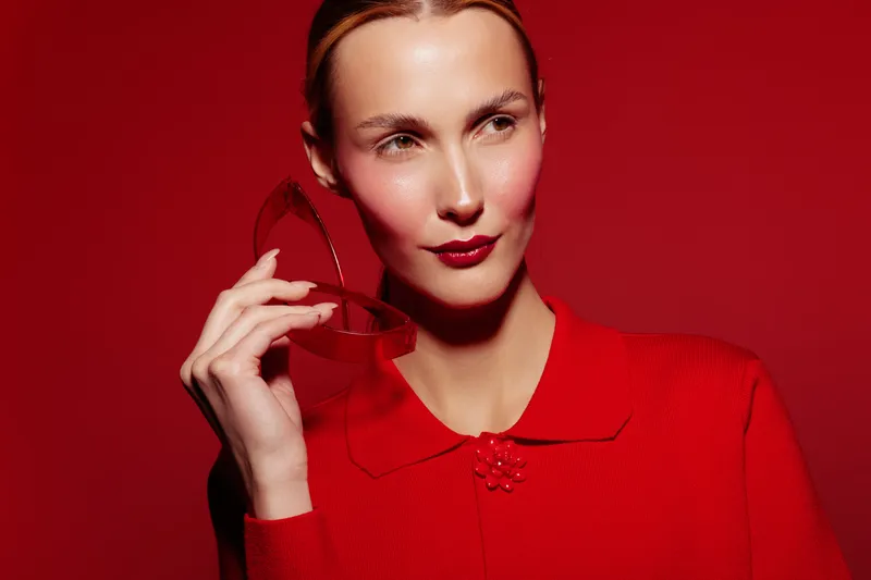 Woman in red sweater holds red sunglasses near face, looking aside on red background. Concept of fashion, style, beauty, and confident female portrait in studio lighting.