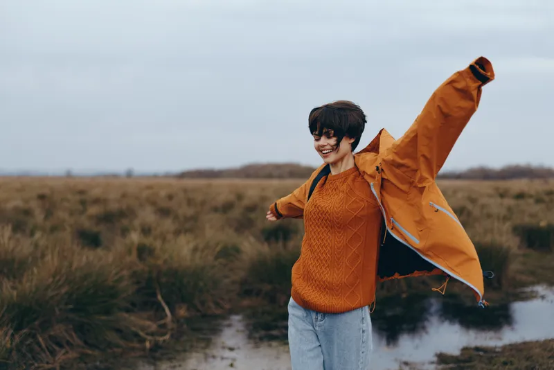 Woman lifestyle outdoors hiking, wearing orange jacket and backpack, smiling with wind in a grassy field near a pond, nature countryside under cloudy sky, travel leisure concept