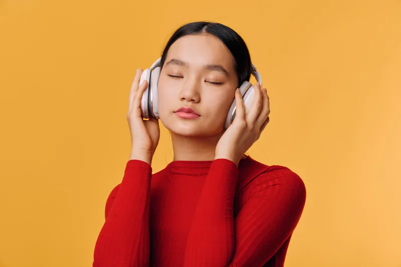 Young woman with closed eyes wearing headphones and red sweater, listening to music or relaxing against orange background. Calm, peaceful and focused female enjoying sound and comfort.