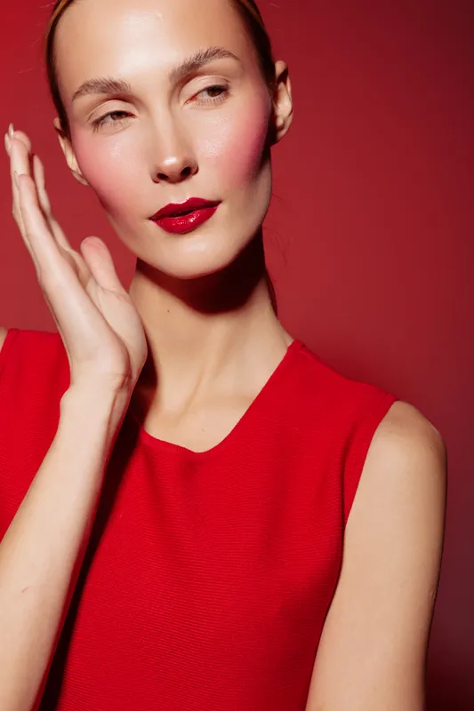 Close-up portrait of a woman with red lipstick and blush wearing a red sleeveless top against a red background. Concept of beauty, fashion, makeup, and elegance.