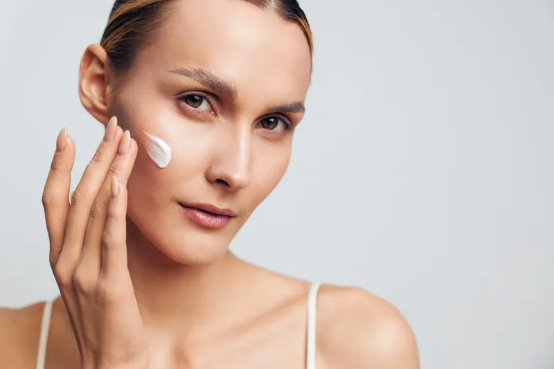 A young woman applies white face cream on her cheek in a close-up portrait with a neutral background. Concept for skincare, beauty routine, and healthy skin care.