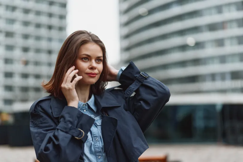 Urban portrait of a woman talking on a smartphone, dressed in a denim jacket, outdoors in front of glass buildings, conveying modern workday communication and confident style