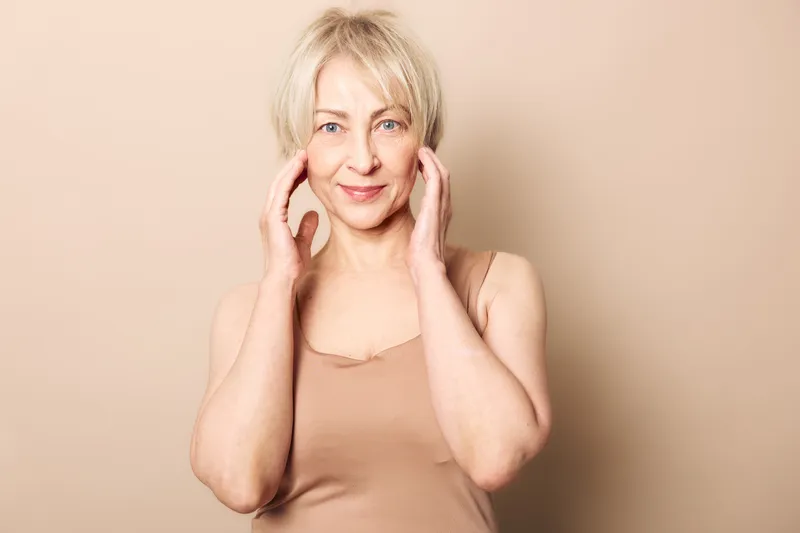 Mature woman with short blonde hair gently touching her face, smiling softly against beige background. Concept of natural beauty, aging gracefully, skin care, and self-confidence.