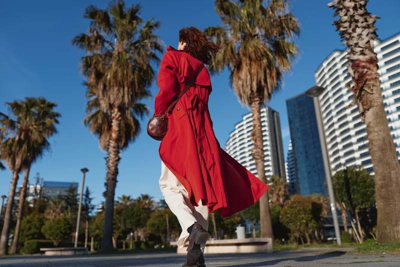 One woman in a bright red trench coat walks briskly through a sunlit urban street lined with palm trees, exuding confident motion and street style.