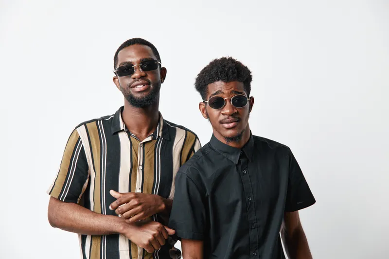 Two young black men with dark skin wearing sunglasses and casual shirts posing together on white background. Friends, style, fashion, confidence, portrait, studio, diversity.