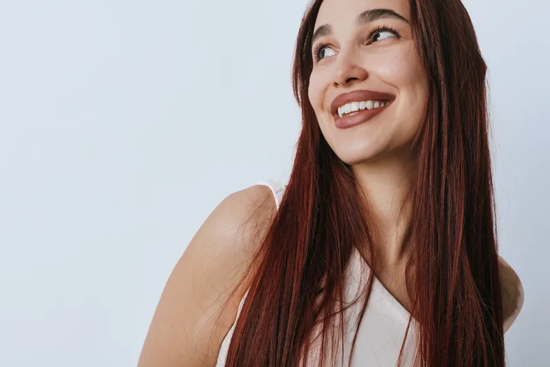 Smiling woman with long red hair looking to side on light background. Portrait of happy female with natural makeup and casual white top in studio with soft lighting.