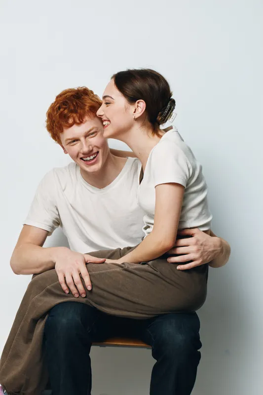 Young couple sitting and laughing together in casual clothes on chair with white background. Man and woman smiling and hugging for friendship and happiness indoors.