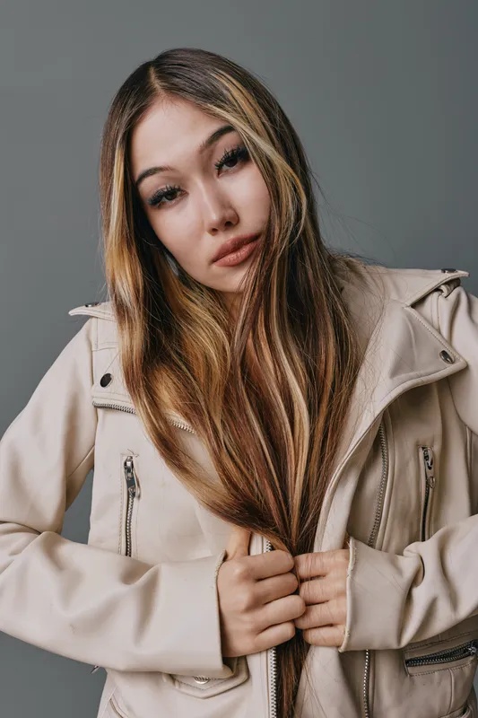 Young woman with long hair wearing beige jacket standing against gray background looking at camera with serious expression and natural makeup in studio portrait.