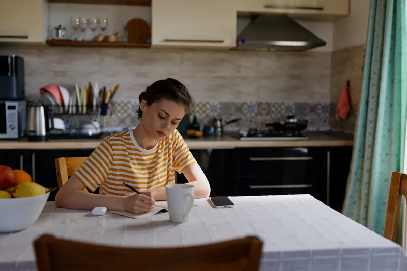 A woman in a striped shirt sits at a kitchen table, focused on writing on paper, with a mug nearby. Bright, cozy home setting conveys everyday study, creativity, and calm afternoon focus.