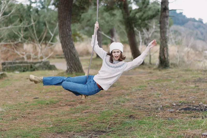Woman on a swing in a park, wearing a cozy sweater and hat, smiling as she soars above the grass with trees in the background, capturing carefree outdoor fun