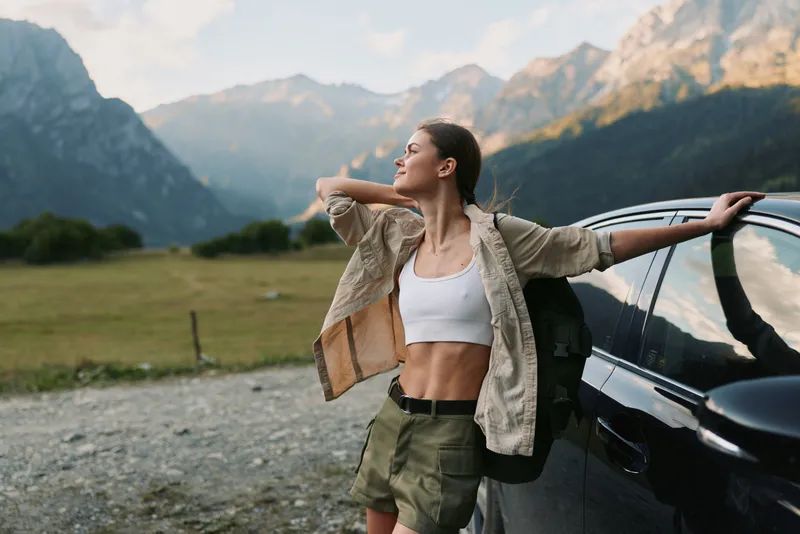 A young woman in casual outdoorwear leans against a dark car beside a mountain valley, arms outstretched, enjoying a moment of freedom after a long hike outdoors.