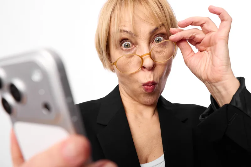 Woman with glasses, surprised expression, smartphone in foreground, hand adjusting gold goggles, studio white background, closeup face portrait, curiosity and astonishment