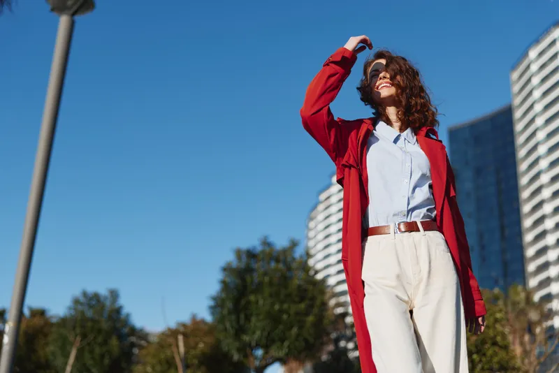 Young woman with curly hair stands outdoors in a red jacket and white shirt, looking up with a bright smile against a clear blue sky amid modern buildings.