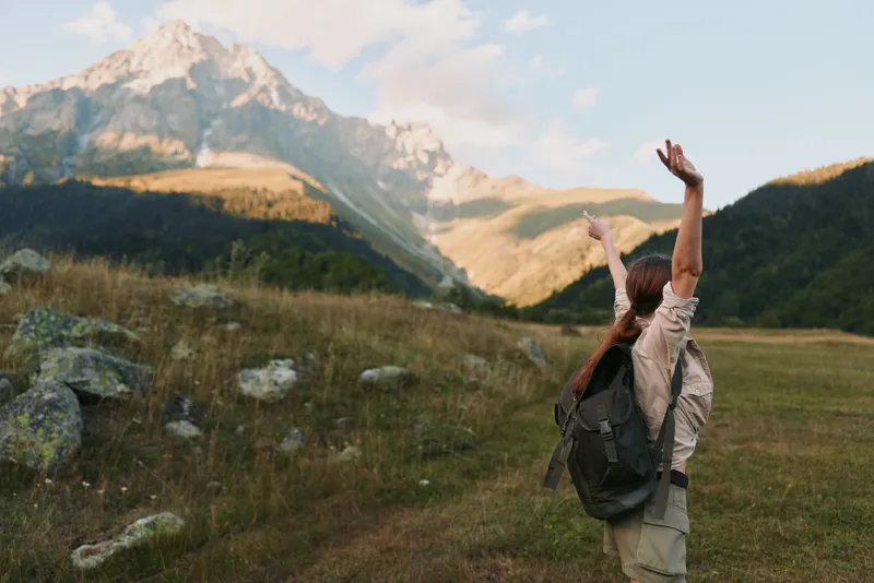 Young woman with a backpack raises her arms in triumph across a sunlit meadow, while rugged mountains loom in the distance and a clear blue sky crowns the landscape.