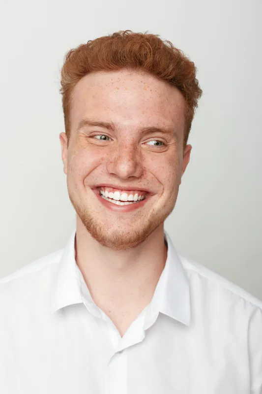 Smiling young man with red hair and freckles looks sideways wearing a white shirt. Bright studio lighting highlights his cheerful expression and natural skin texture on grey background.