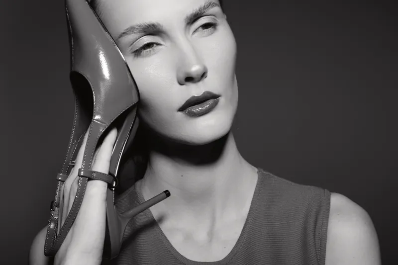 White woman with pale skin holds high heel shoe to her face in black and white close-up. Concept of fashion, beauty, and artistic portrait.