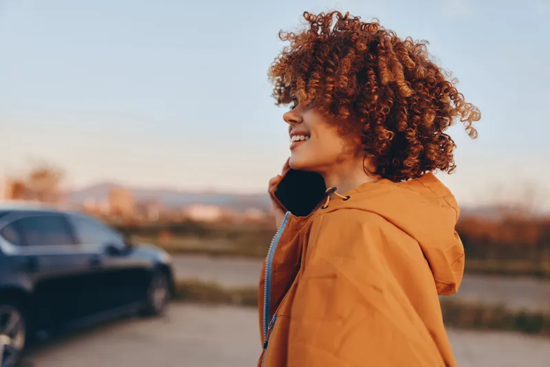 Woman lifestyle profile portrait, curly hair, smiling outdoors, wearing orange jacket, sunset light, soft background with shallow depth of field, parked cars nearby, casual street travel moment