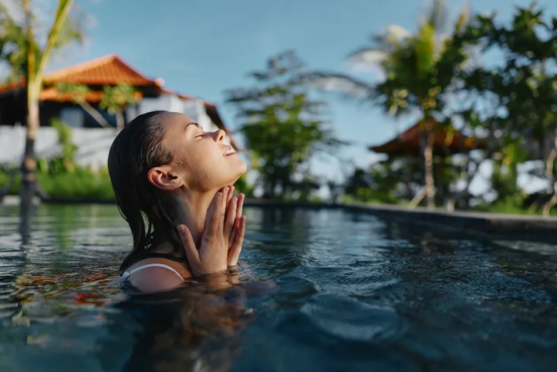 Relaxed woman in a sunny pool with eyes closed, hands at her neck, embracing calm, clear water, tropical resort backdrop, wellness lifestyle, tranquil mood, soft natural light