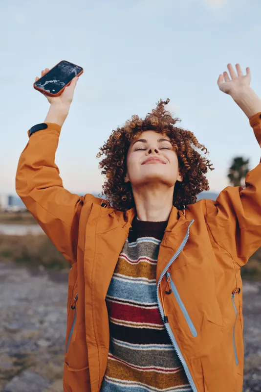 Woman lifestyle curly hair outdoors smartphone jacket joy, orange jacket striped sweater, arms raised with smartphone, smiling face in bright light, blurred street background, sky