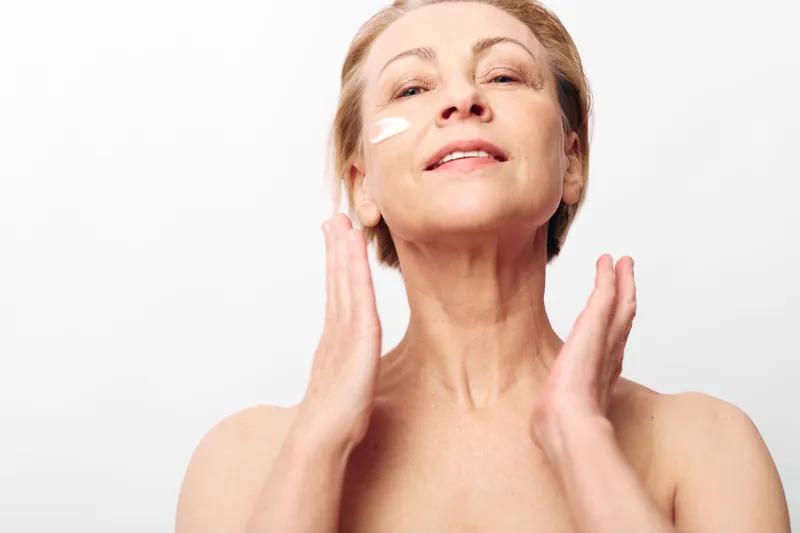 Mature woman applies face cream on clean skin with gentle hands. Soft natural skincare routine, beauty care concept on white background. Healthy skin and self-care moment.