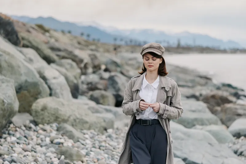 Young woman in casual outfit walks on rocky beach near calm sea with mountains in background. Overcast sky and muted colors create peaceful, contemplative mood in nature setting.