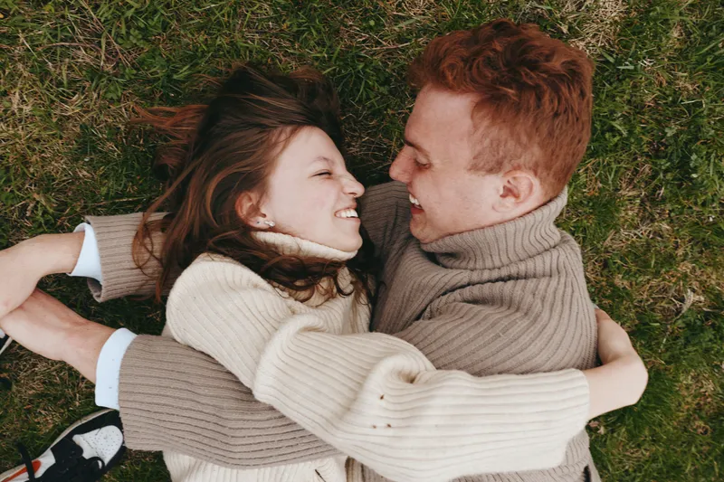 Romantic couple lying on grass outdoors, smiling and embracing, cozy sweaters, warm mood, hands intertwined, candid lifestyle moment, close connection, natural light, peaceful atmosphere.
