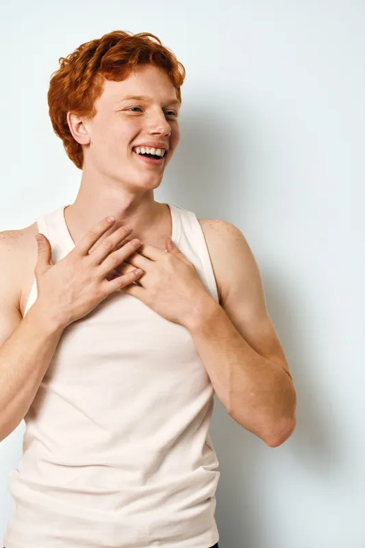 Young man with red hair smiling and touching chest in white tank top on plain background. Casual portrait of happy person in bright studio lighting with joyful expression.