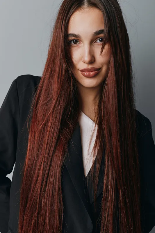 Woman with long straight brown hair wearing black blazer and white shirt posing against gray background. Portrait of confident female model with natural makeup and casual hairstyle indoors.
