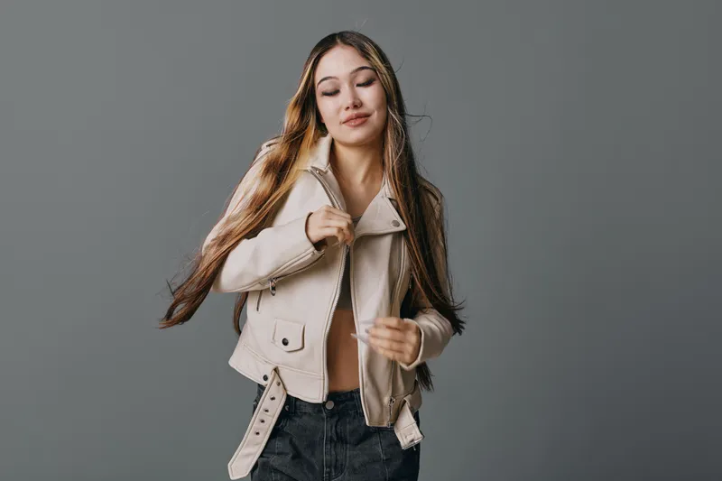 Young woman with long hair wearing beige jacket and black pants posing on gray background, fashion model with casual style and natural makeup for portrait photography.