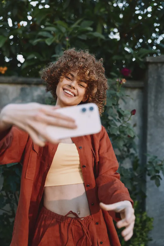 Joyful woman with curly hair in a burnt orange outfit takes a selfie outdoors, smiling at the camera, garden background, candid casual lifestyle portrait