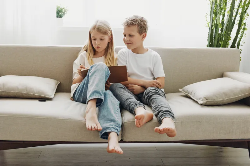 Two children sitting barefoot on beige sofa playing with tablet together in bright modern living room with plants and cushions.