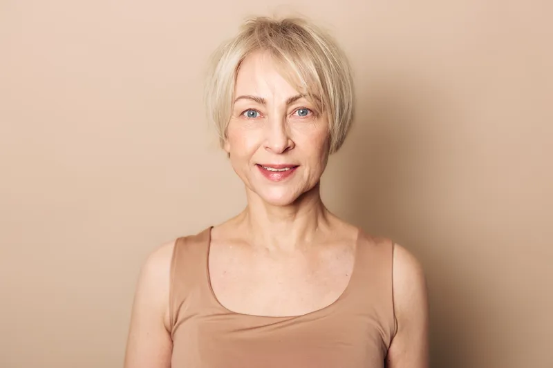 Portrait of a mature woman with short blonde hair smiling gently against beige background. Natural expression highlights beauty, confidence, and healthy skin in soft lighting.