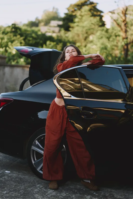 Smiling woman wearing red jumpsuit leans on a sleek car with an open trunk outdoors, showcasing casual lifestyle and street fashion, natural light creating a warm, confident mood