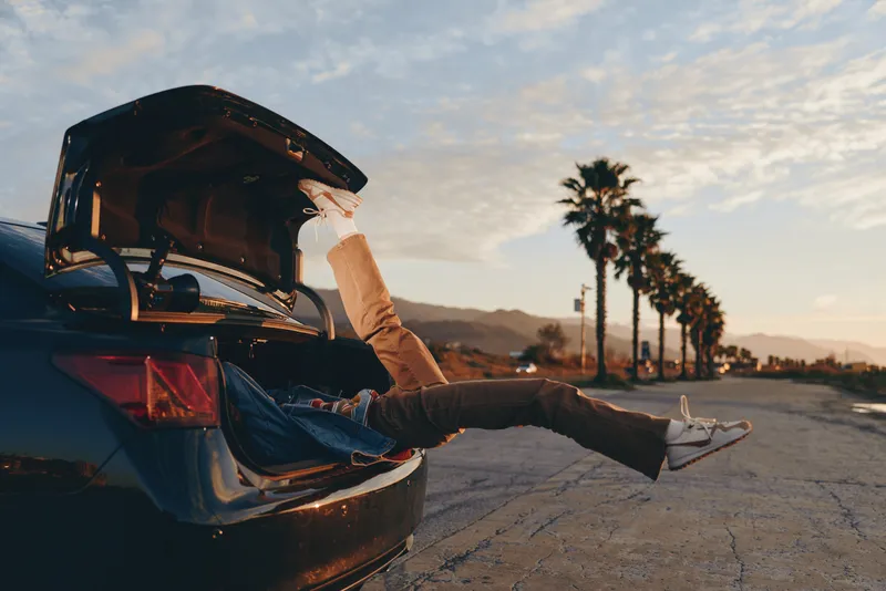 Lifestyle photo, man relaxing by the open car trunk, legs on the road, sunset light, palm trees ahead, outdoors travel mood, casual jeans and sneakers, calm cinematic horizon view warm air