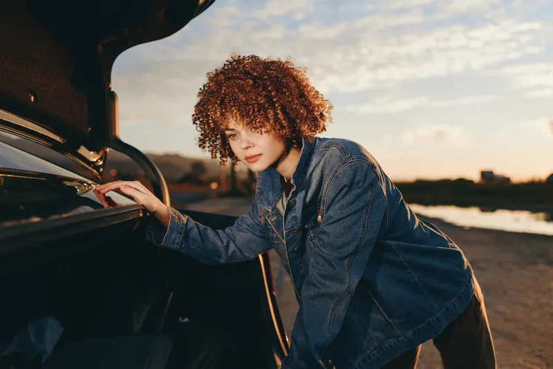 Woman lifestyle scene, curlyhair woman in blue denim jacket at a car, hand on open hood, golden sunset sky, outdoors by road, urban city parking
