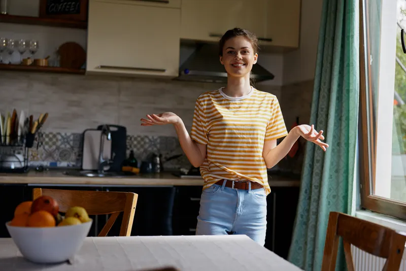 Smiling woman in a striped shirt stands in a bright kitchen, gesturing casually with open hands. The cozy home setting conveys everyday life, comfort, and relaxed wellbeing and vitality.