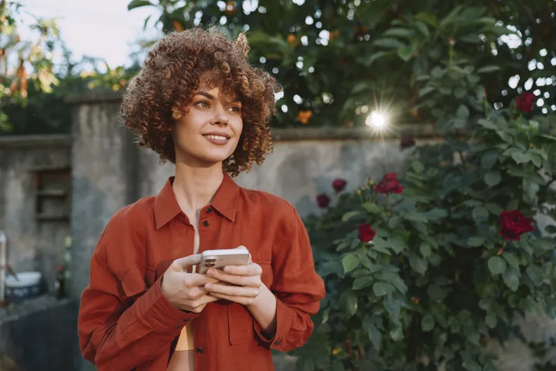 A woman with curly hair in a red shirt stands outdoors, holding a smartphone and smiling. Sunny garden setting with flowers in the background. A lifestyle portrait about everyday joy