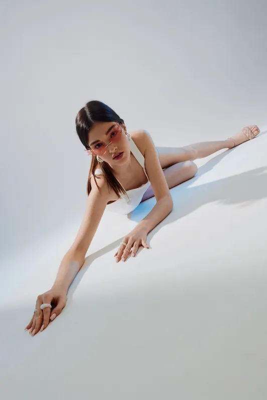 Young woman in white fashion outfit poses on floor with stretched arms in bright studio. Concept of beauty, fashion, elegance, and modern style.