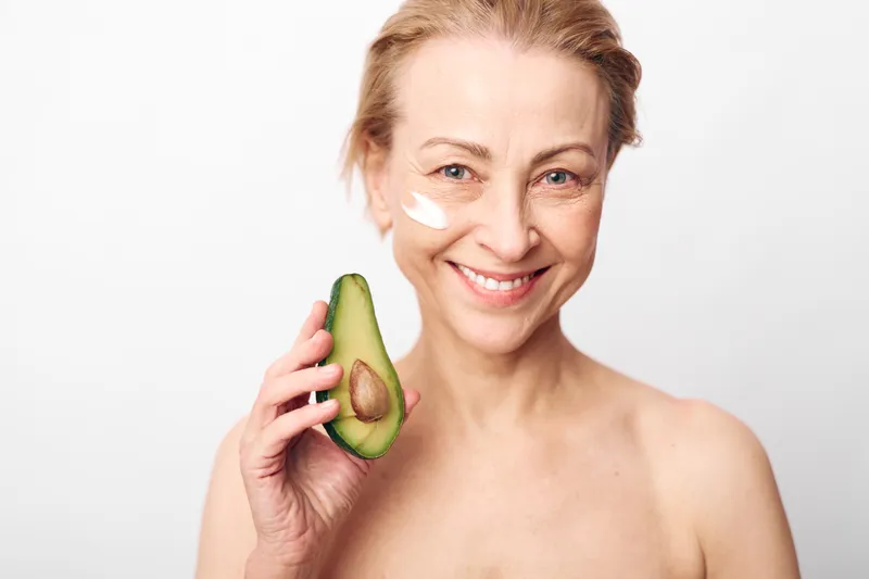 Smiling mature woman holding half of avocado with natural cream on face, isolated on white background. Concept of skincare, healthy lifestyle, and natural beauty care routine.
