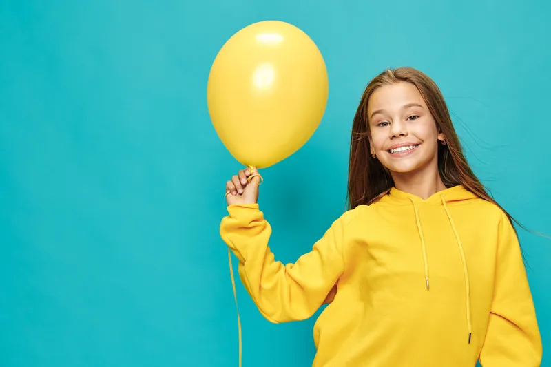 Girl smiling and holding yellow balloon wearing yellow hoodie on blue background. Child with long hair and cheerful expression in casual clothes for celebration or fun activity.