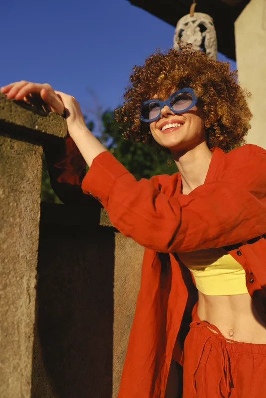 A joyful woman with curly hair in a red jacket and yellow top leans on a sunny outdoor wall, smiles while wearing blue sunglasses. A bright, lifestyle moment celebrating creativity and warmth