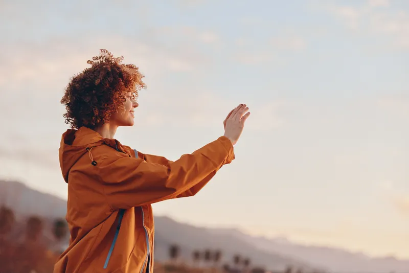 Woman lifestyle portrait outdoors at sunrise, curly hair and jacket, waving gesture toward mountain landscape, warm sunshine light, calm travel mood, countryside horizon, soft background