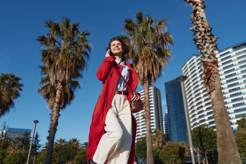 Smiling woman in a red coat walking outdoors in a sunny urban setting with palm trees and tall buildings. Fashion focus on style, confidence, and street vibe.